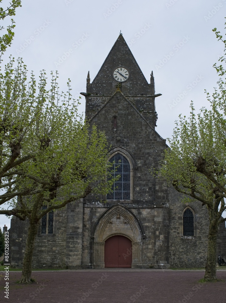 Fototapeta premium Sainte-Mere-Eglise, France - Apr 19, 2024: Sainte-Mere-Eglise church. People walking in Sainte-Mere-Eglise. Streets and buildings. Lifestyle in the urban area. Cloudy spring day. Selective focus