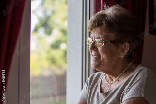 Pensive thoughtful lonely grandma standing alone at home looking through window waiting. Senior smiling woman waiting for visitors by the window.