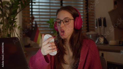 Close up, a young confident brunette girl with glasses in red wireless headphones and a sweater studies a foreign language German and looks at herself with the help of a mirror while pronouncing words photo