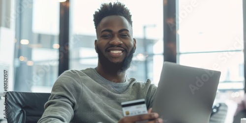 Young joyful black guy working alone with a credit card and laptop. A joyful guy business professional using a debit card to buy something online at a desk.