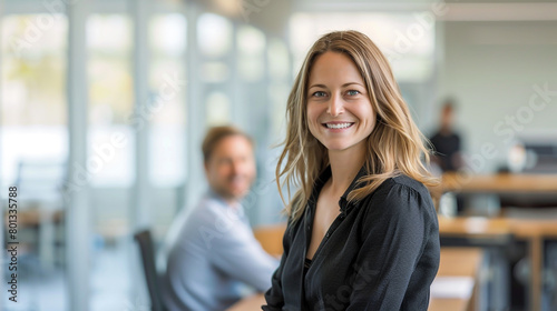 In an office, a smiling businesswoman stands while a man sits behind a desk, observing her with a blurred background. Free Download In an office, a smiling businesswoman stands while a man sits behind a desk, observing her with a blurred background. - GFXLoad