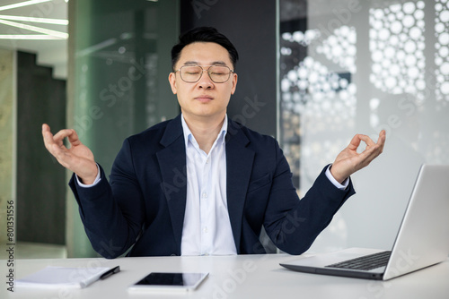 A professional businessman in a modern office meditating at his desk. He practices mindfulness to manage stress and enhance focus amid a busy work environment.