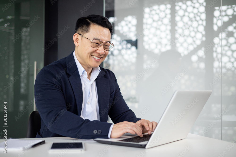 Joyful Asian businessman smiling while typing on his laptop in a contemporary office setting, reflecting success, productivity, and happiness at work. Joyful Asian businessman smiling while typing on his laptop in a contemporary office setting, reflecting success, productivity, and happiness at work.