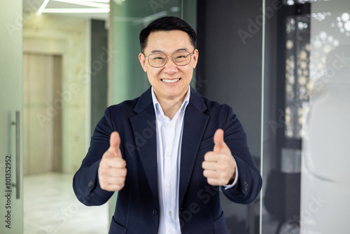 A professional Asian businessman in a navy suit stands in a modern office, smiling and giving a thumbs up, exuding confidence and success.