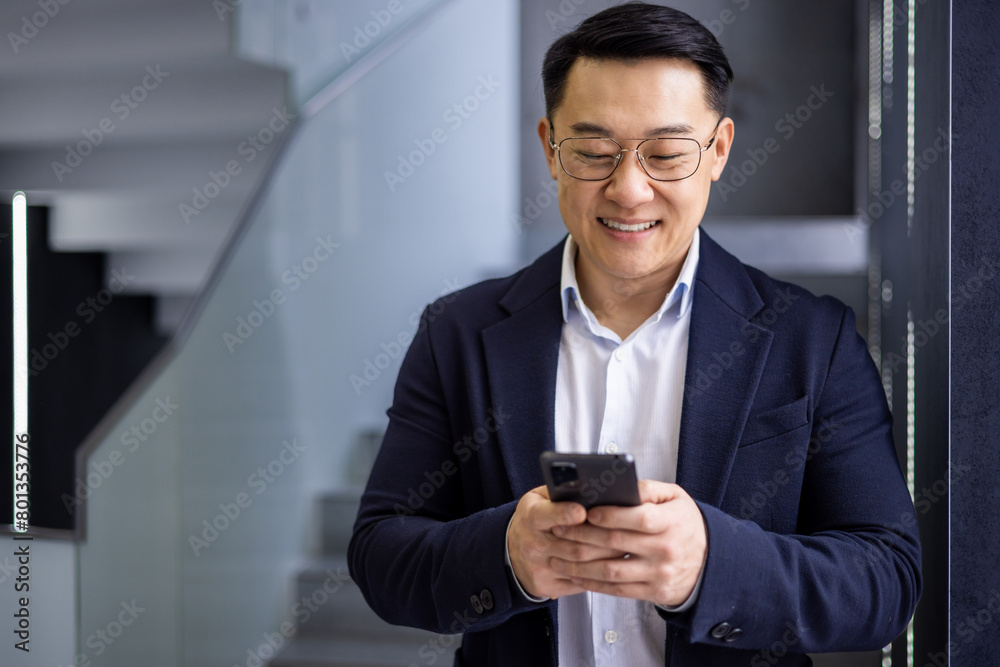 A cheerful Asian businessman in a dark suit is using his smartphone, standing in a stylish modern office setting, looking engaged and happy.