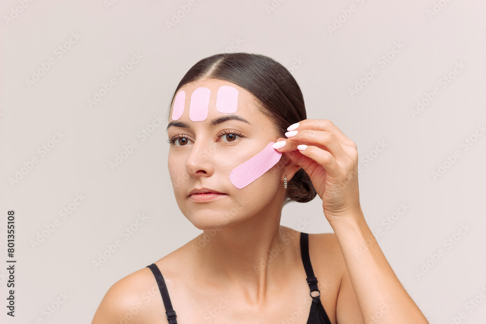A young brunette Caucasian woman puts a pink kinesiotaping tape on her face to prevent the appearance of wrinkles, tighten the oval of her face and eliminate swelling. Facial care. Slowing down aging A young brunette Caucasian woman puts a pink kinesiotaping tape on her face to prevent the appearance of wrinkles, tighten the oval of her face and eliminate swelling. Facial care. Slowing down aging