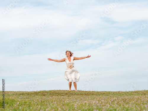Quiberon, France, April 17, 2024. Woman in white dress and long red hair, happy runs to greet, hands spread wide against the blue sky, green grass nature, happiness freedom