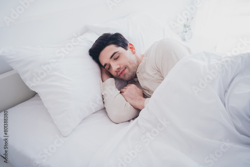 Photo of young attractive man enjoying sleepwear lying in soft comfy bed white room interior inside