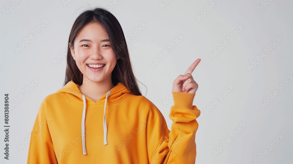 Young Asian woman in a yellow hoodie smiling and pointing to the side on a light background. Young Asian woman in a yellow hoodie smiling and pointing to the side on a light background.