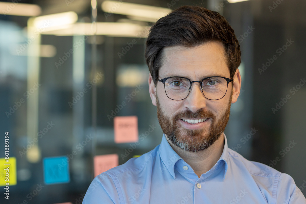 Obraz premium A professional portrait of a confident, smiling businessman wearing glasses and a blue shirt, shot in a bright office environment with sticky notes in the background.