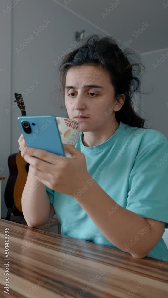 Young woman drinks tea checking social networks on smartphone. Sleepy housewife uses electronic device sitting at wooden table in morning Vertical footage. Young woman drinks tea checking social networks on smartphone. Sleepy housewife uses electronic device sitting at wooden table in morning Vertical footage.