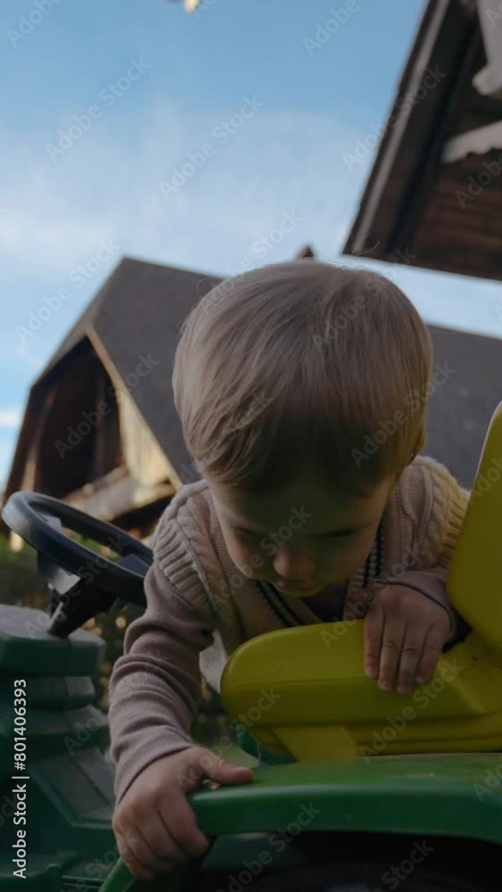 Cute blond toddler tries to get on small tractor on green lawn in yard of country house. Active child enjoys examining vehicle toy slow motion Vertical footage. Cute blond toddler tries to get on small tractor on green lawn in yard of country house. Active child enjoys examining vehicle toy slow motion Vertical footage.