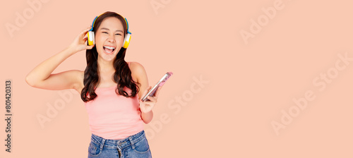Happy young asian woman laughing while listening to music on yellow wireless headphones and holding a smartphone, isolated on a peach background and copy space
