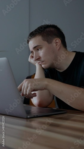 Depressed man tries hard to continue work on laptop sitting at wooden table in room. Male freelancer pushes buttons on keyboard of device feeling bad Vertical footage.