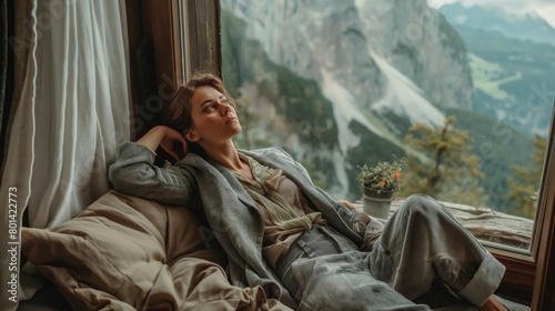 A woman resting comfortably, dressed in a white outfit and gray blazer, with a scenic mountain view in the background. An indoor portrait with natural lighting, representing comfort workation