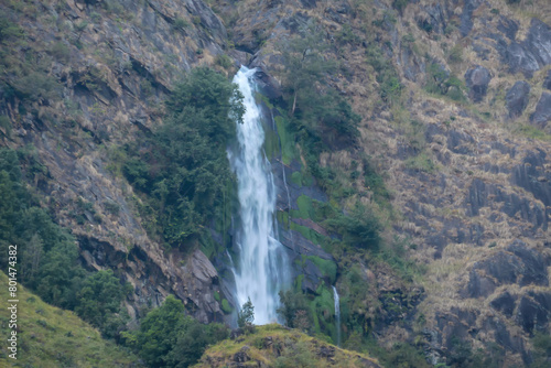 A tall waterfall in Himalayas, along Annapurna Circuit Trek in Nepal. The water free falls from a high distance. The slopes of the mountain are overgrown with lush green plants. photo