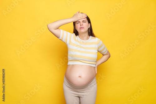 Unhealthy Caucasian pregnant woman with bare belly wearing casual top isolated over yellow background feeling fatigue touching her painful head having high temperature during pregnancy photo