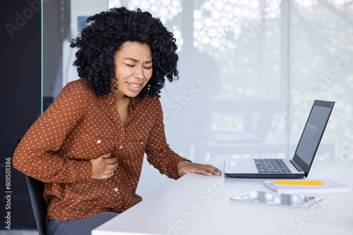 A young African American woman feeling unwell with a stomach ache as she works on her laptop in a modern office setting.