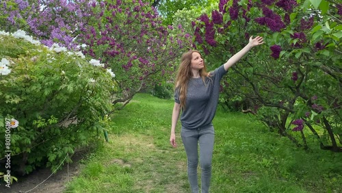 A girl walks along a blooming lilac and touches a flower petal with her hand. A walk in the park. The girl runs her hand over the red lilac flowers, and then bends the branch and smells it.