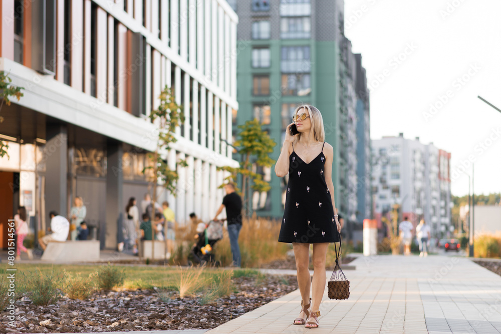 Young woman in a black dress, talking on a cell phone, walking on a city street in the summertime Young woman in a black dress, talking on a cell phone, walking on a city street in the summertime