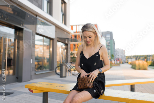 Young Caucasian blonde woman sits on a bench looking in her bag for her phone or money