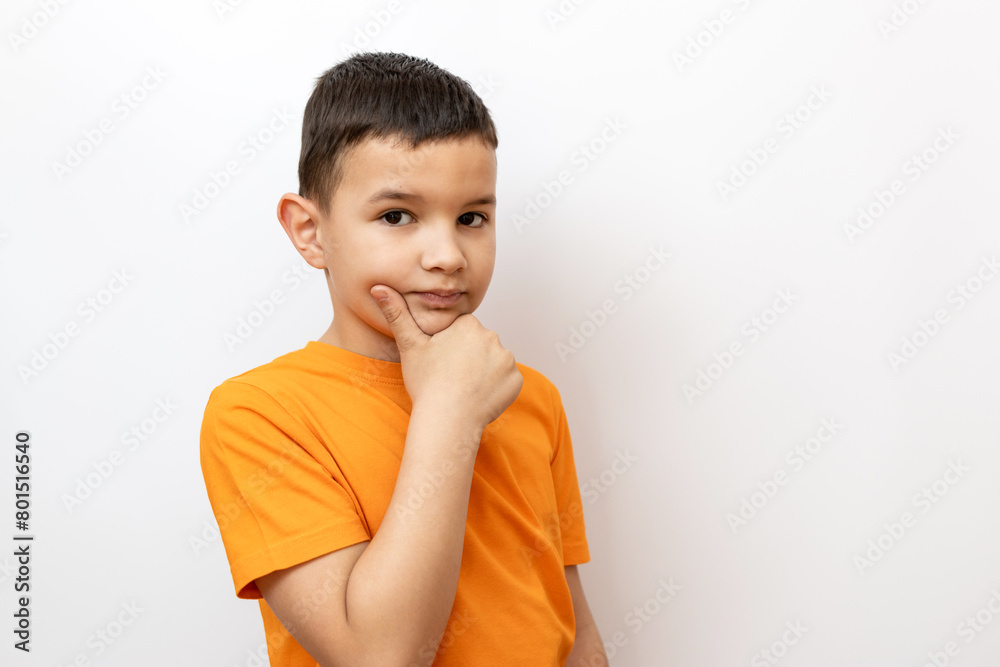 A boy in an orange T-shirt on a white background holds his hand on his chin.