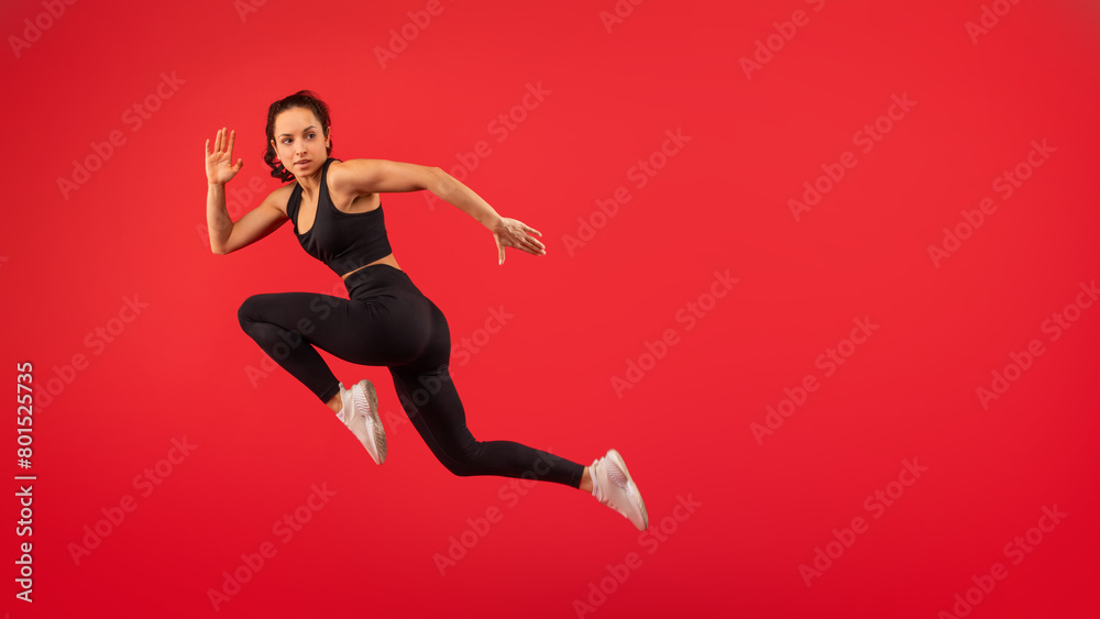 A woman wearing a black top and black leggings is captured mid-jump in the air. Her arms are outstretched and her legs are bent, showcasing her athleticism and agility as she defies gravity.