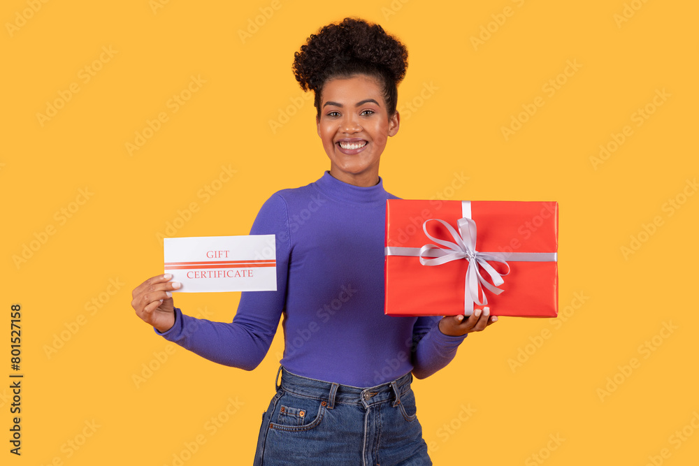 Hispanic young woman standing while holding two gift cards and a present in her hands, yellow background Hispanic young woman standing while holding two gift cards and a present in her hands, yellow background