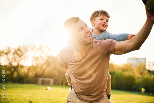 On shoulders, with soccer ball. Happy father with son are having fun on the field at summertime