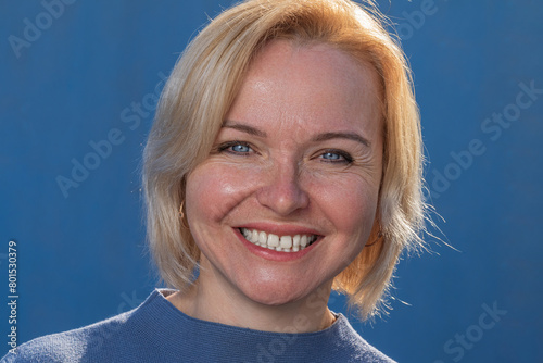 Adult and happy blonde close-up smiles broadly on a blue background in a blue sweater photo