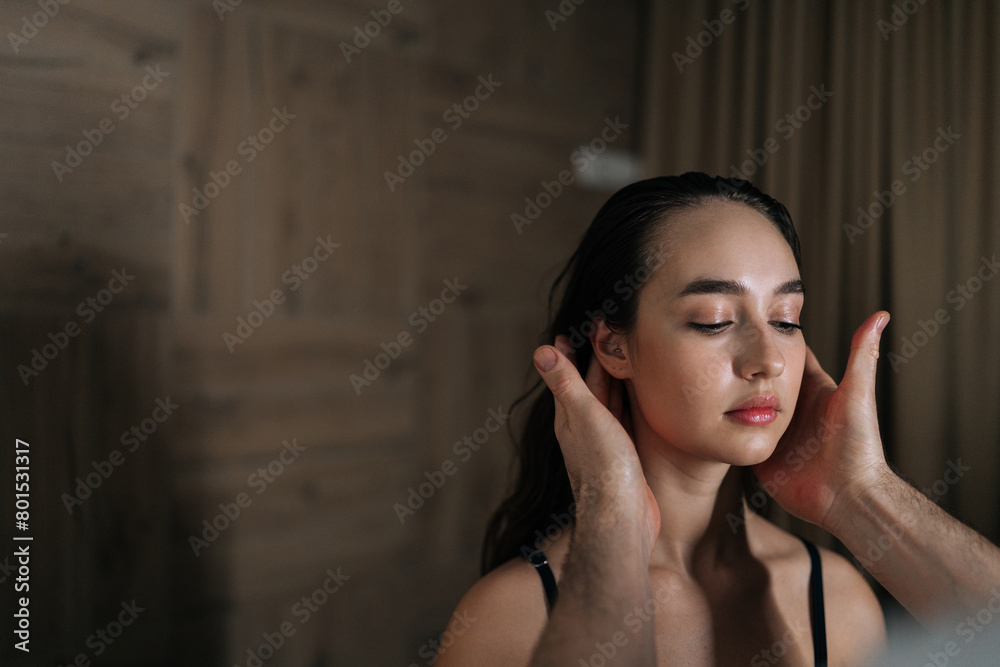 Serene young woman with closed eyes relaxed while masseur massaging ears in wellness center with dark interior. Professional masseur male touching auricle of beautiful female. Concept of spa treatment Serene young woman with closed eyes relaxed while masseur massaging ears in wellness center with dark interior. Professional masseur male touching auricle of beautiful female. Concept of spa treatment