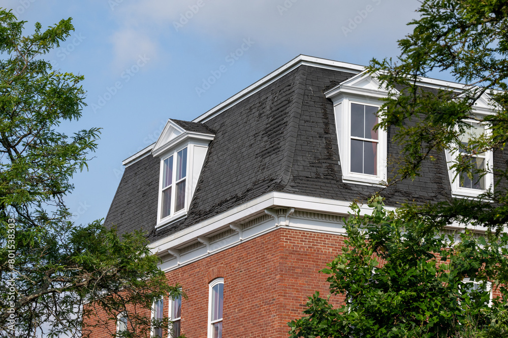 The exterior wall of a tall brown brick building with multiple rows of double hung windows. The windows have white trim. The hip roof is made of black felt. The windows in the roof are peaked gable. 