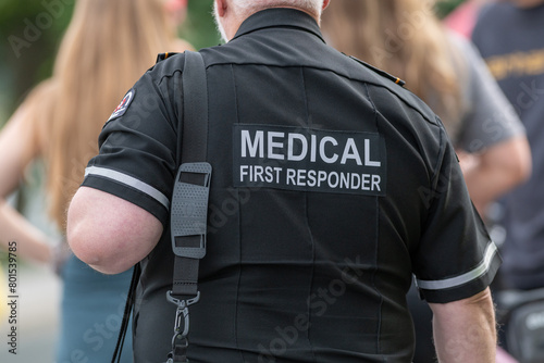 A close-up of a Caucasian male emergency health medical first responder or paramedic wearing a black uniform with grey letters. The ambulance attendant is wearing a short-sleeve shirt with lettering.