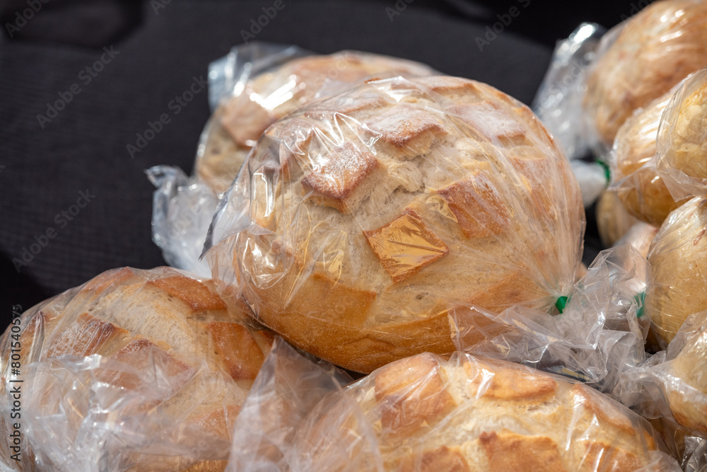 Multiple bags of fresh sourdough bread loaves. The golden crusted rustic bread has been scored on top and baked in a Dutch oven. The unsliced white and wheat flour loaves are in transparent bags.