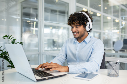 A cheerful young man with curly hair wears headphones while focusing on work using a laptop in a modern office environment, showcasing productivity and concentration.