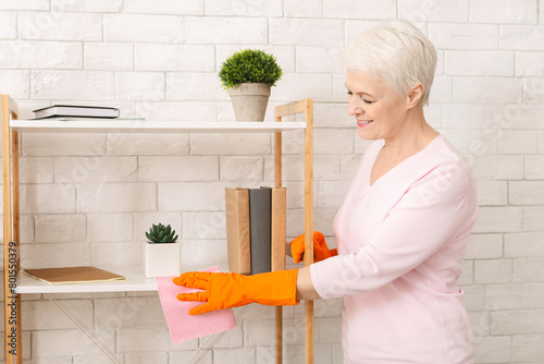 Attractive elderly woman is standing in a room, leaning forward to clean a shelf with a yellow sponge. She is focused on removing dust and grime from the surface.