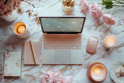 Laptop is on a table with a pink notebook, a pen, a mouse, and a cup of coffee, flowers and candles, creating a cozy atmosphere