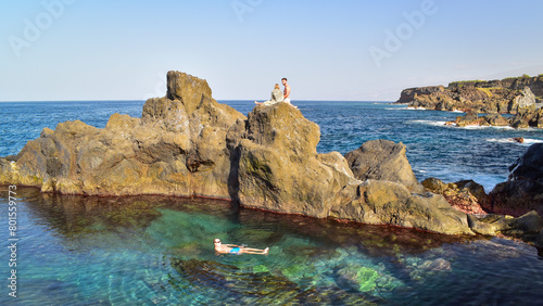 Man floating in clear blue water, couple sunbathing on rocks. Tenerife North Coast, Canary Islands Spain.