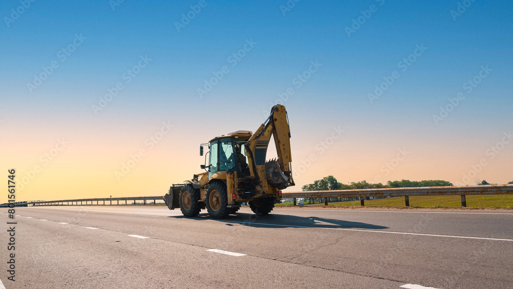 Yellow backhoe loader traveling on highway under clear blue sky signifying ongoing construction work. Open road and sunny day provide perfect setting for heavy machinery operation Yellow backhoe loader traveling on highway under clear blue sky signifying ongoing construction work. Open road and sunny day provide perfect setting for heavy machinery operation