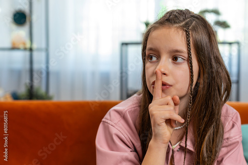 Shh be quiet please. Portrait of preteen girl child presses index finger to lips makes silence hush gesture sign do not tells gossip secret. Little cute teenage female sitting on sofa in living room.