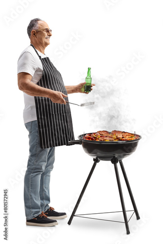 Mature man wearing apron holding a bottle of beer and grilling meat on a barbecue