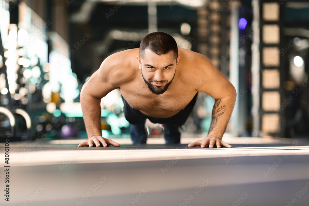 Shirtless man is performing push ups in a gym, his muscles strained as he lowers and raises his body. The gym equipment and other people working out in the background add to the active atmosphere. Shirtless man is performing push ups in a gym, his muscles strained as he lowers and raises his body. The gym equipment and other people working out in the background add to the active atmosphere.