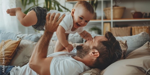 A handsome young father is lying on the sofa, holding his one-year-old baby boy in midair and lifting him up with both hands to play photo