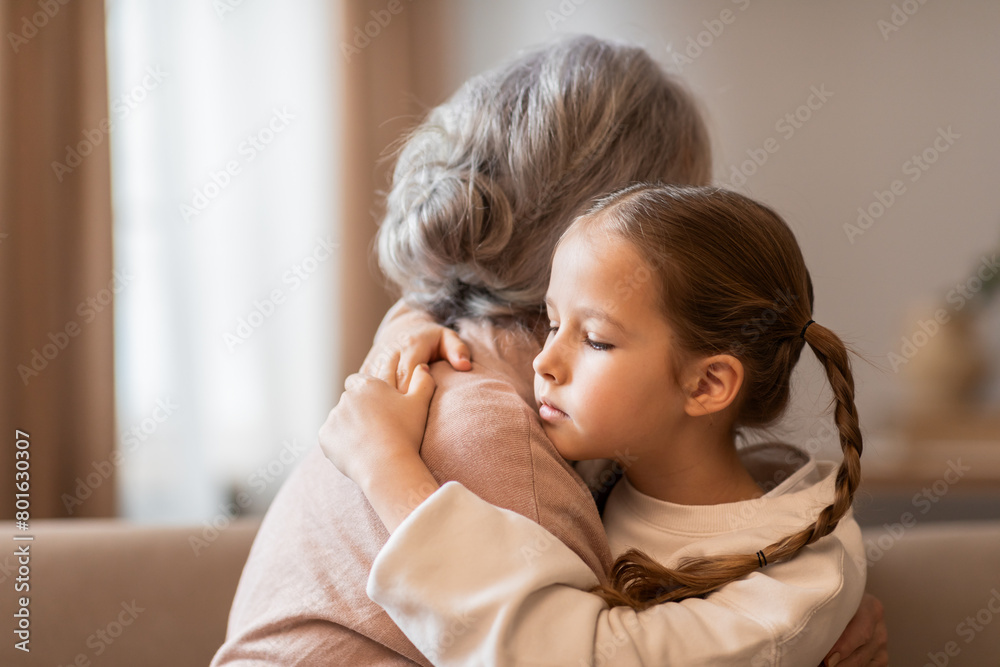A young girl embraces a senior woman warmly as they sit together on a couch in a cozy living room. The child arms are wrapped around the older woman A young girl embraces a senior woman warmly as they sit together on a couch in a cozy living room. The child arms are wrapped around the older woman