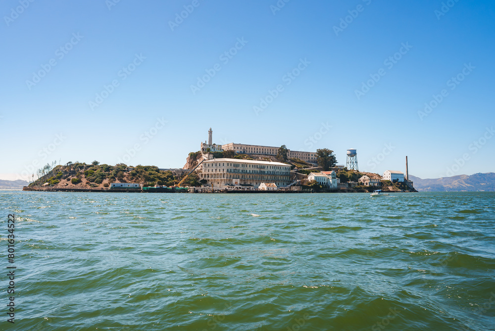 Alcatraz Island view under clear sunny skies, featuring Alcatraz Federal Penitentiary structures from the bay in San Francisco Bay. Notable landmarks visible.