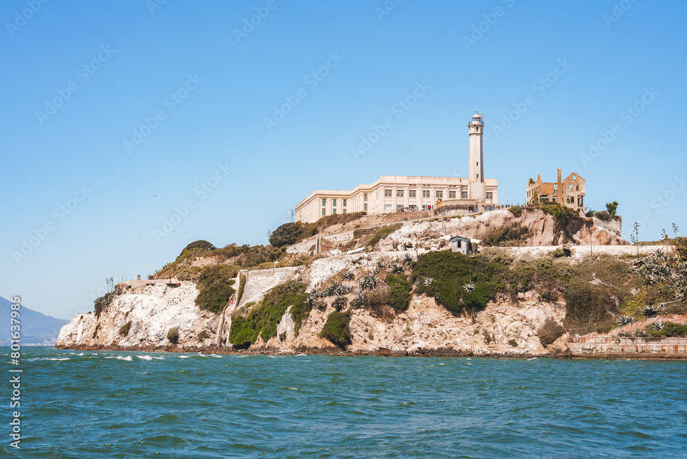 Alcatraz Island view from water in San Francisco Bay, California, USA. Rugged coastline, former prison, iconic lighthouse, clear sky, calm water, sparse vegetation, historical landmark.