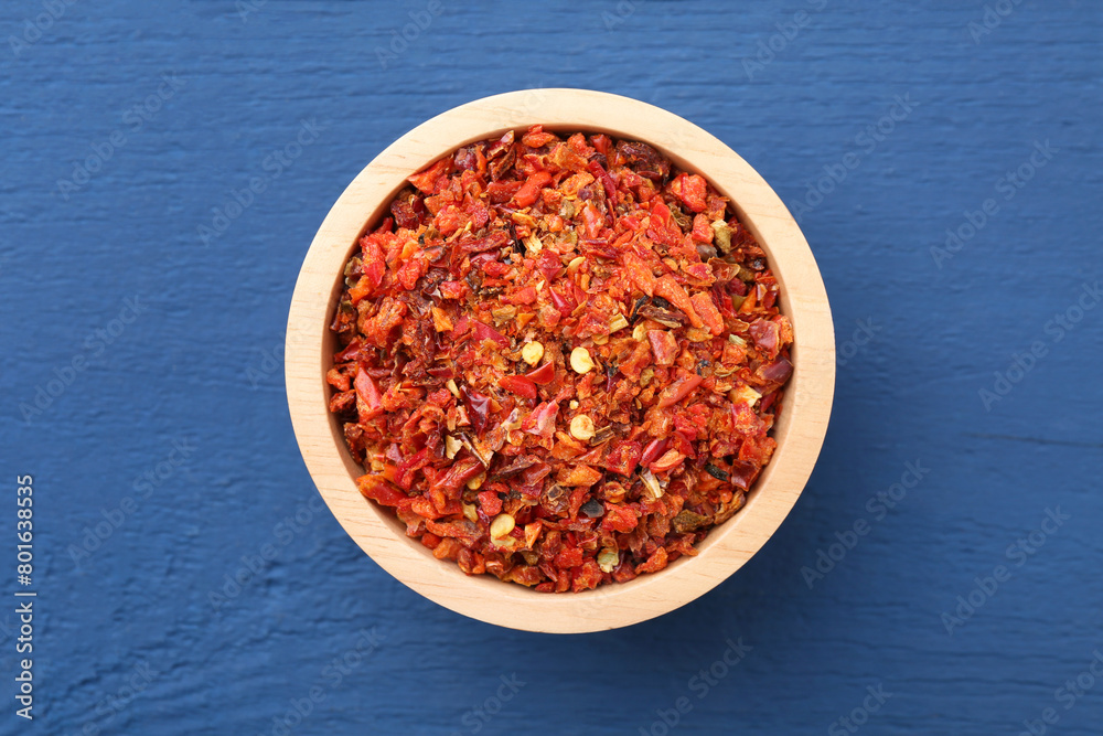 Chili pepper flakes in bowl on blue wooden table, top view