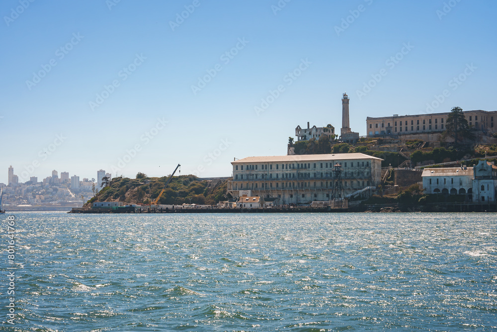 View of Alcatraz Island in San Francisco Bay, California, USA on a clear day. Iconic buildings and lighthouse against a blue sky, with cityscape in the background.