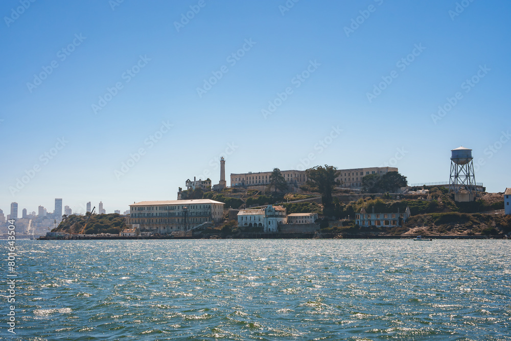 View of rugged Alcatraz Island in San Francisco Bay, California, USA. Shows main cellhouse, guard tower, choppy waters, and city skyline in background.