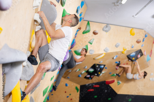 European man is mastering climbing on training wall in gym, side view. He holds on tightly to ledges and strives for top of bouldering route